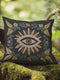 A black woven throw pillow featuring intricate embroidery of an Evil Eye motif in gold, ivory, and green, surrounded by detailed botanical flourishes.